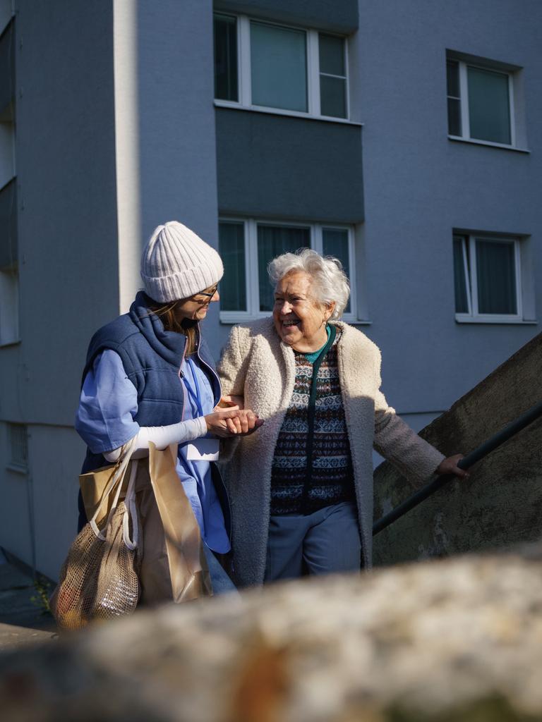 Eine Frau begleitet eine Seniorin am Arm die Stufen vor einem Wohnhaus hinauf. Auf dem Arm trägt sie Papiertaschen mit Einkäufen.

