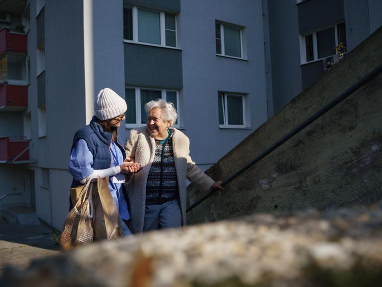 Eine Frau begleitet eine Seniorin am Arm die Stufen vor einem Wohnhaus hinauf. Auf dem Arm trägt sie Papiertaschen mit Einkäufen.

