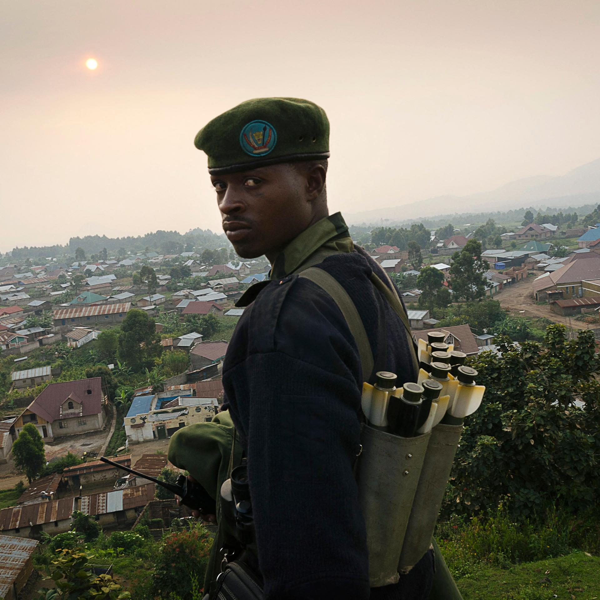 Ein uniformierter Kämpfer schaut in die Kamera. Er steht auf einem Hügel, hinter ihm sind die Dächer einer Ortschaft zu sehen.