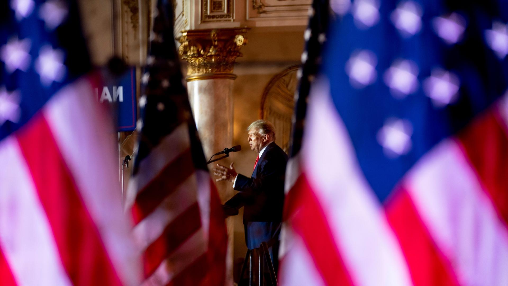 Donald Trump steht im Hintergrund an einem Rednerpult und spricht. Im Vordergrund, in der Unschärfe, ist links und rechts die amerikanische Flagge zu sehen.