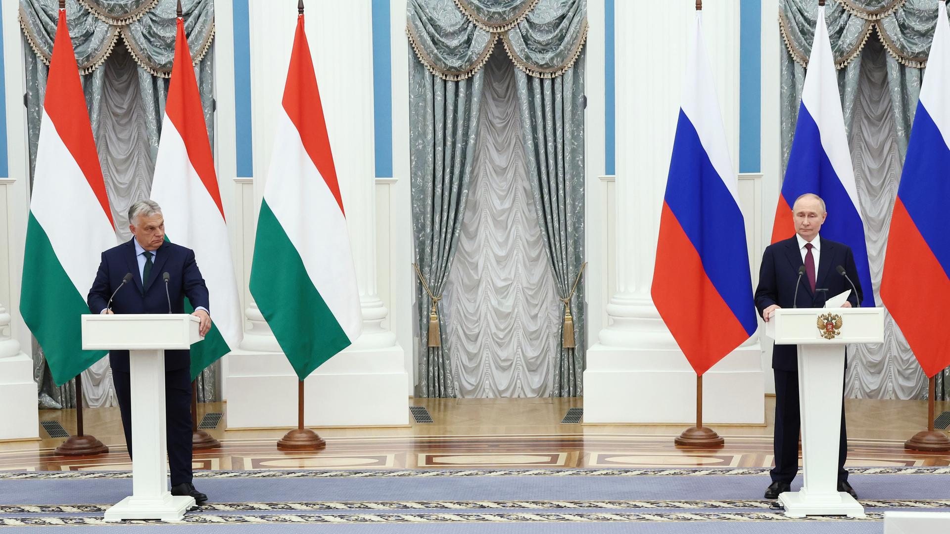 Viktor Orban und Wladimir Putin stehen an zwei etwas weiter auseinanderstehenden Pulten und geben ein Presse-Statement ab. Im Hintergrund sind vor einer prunkvollen Wand des Kreml die Flaggen Ungarns und Russlands zu sehen.
