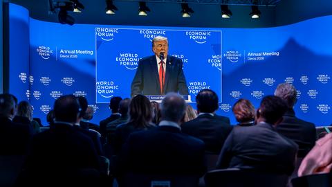 US-Präsident Donald Trump ist auf einer Großbildleinwand zu sehen, während er eine Rede hält. Im Vordergrund einige Zuhörerinnen und Zuhörer.