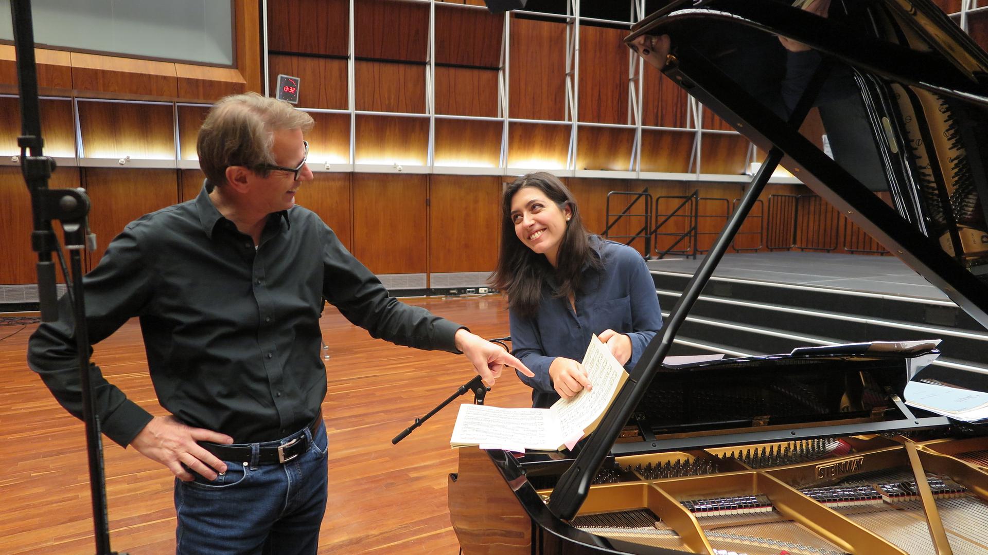 Ein Mann mit Brille und eine Frau mit dunklen Haaren stehen an einem Flügel und zeigen lächelnd auf eine Notenpartitur. Ein Mann mit Brille und eine Frau mit dunklen Haaren stehen an einem Flügel und zeigen lächelnd auf eine Notenpartitur.