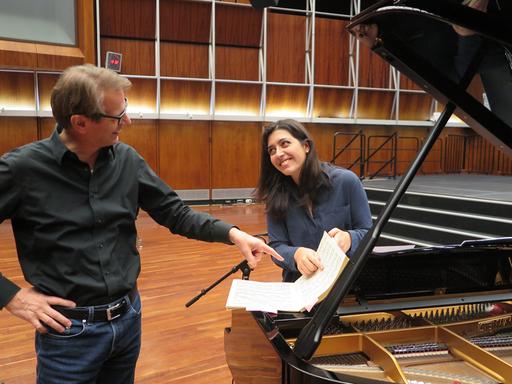 Ein Mann mit Brille und eine Frau mit dunklen Haaren stehen an einem Flügel und zeigen lächelnd auf eine Notenpartitur.