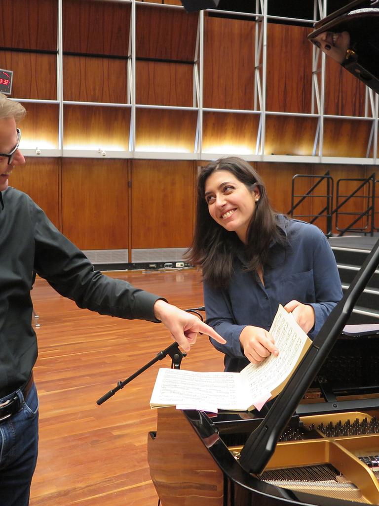 Ein Mann mit Brille und eine Frau mit dunklen Haaren stehen an einem Flügel und zeigen lächelnd auf eine Notenpartitur.