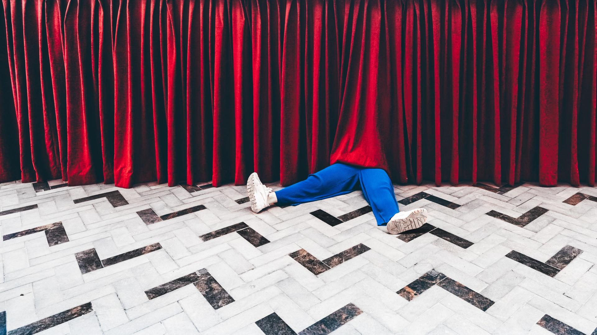 Beine einer Person in blauen Hosen schauen unter einem Theatervorhang heraus.