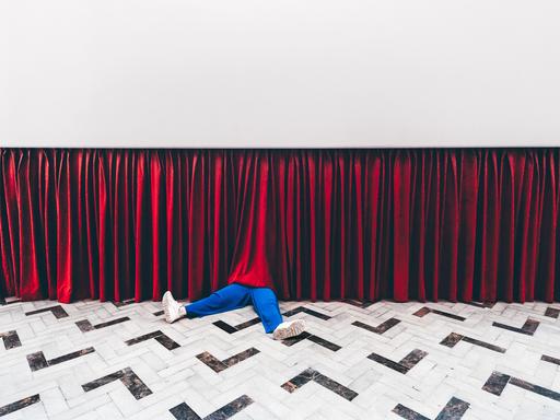 Beine einer Person in blauen Hosen schauen unter einem Theatervorhang heraus.