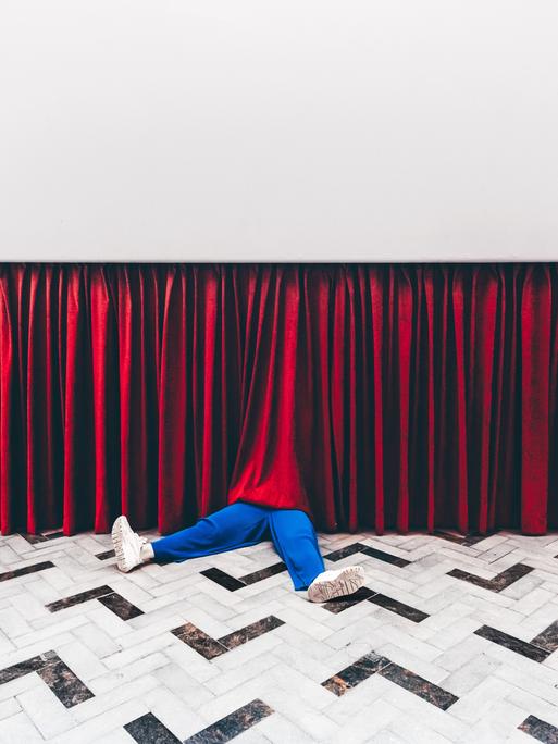 Beine einer Person in blauen Hosen schauen unter einem Theatervorhang heraus.