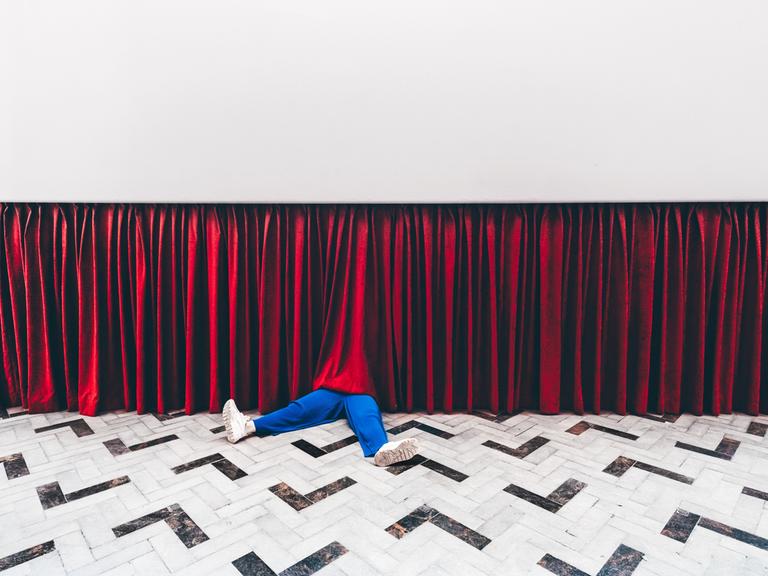 Beine einer Person in blauen Hosen schauen unter einem Theatervorhang heraus.