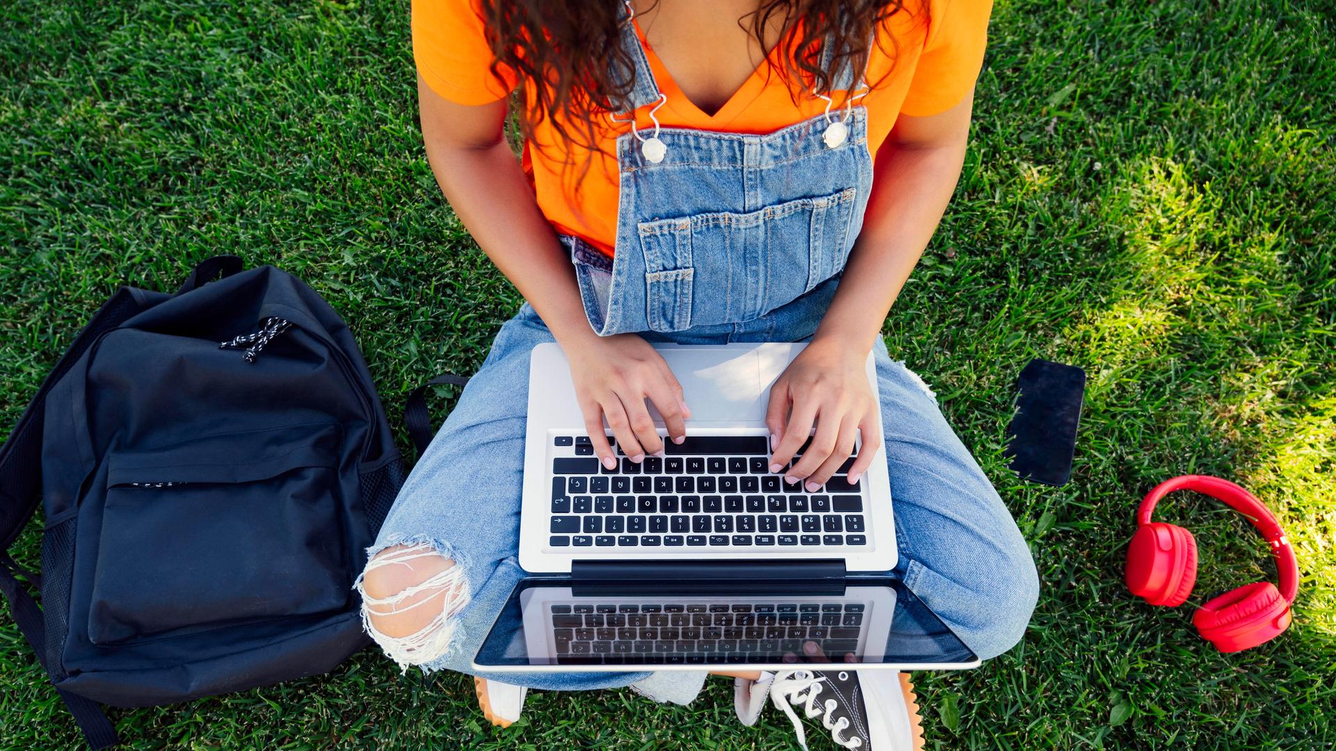 Eine junge Frau sitzt im Gras und tippt etwas in ihren Laptop. 