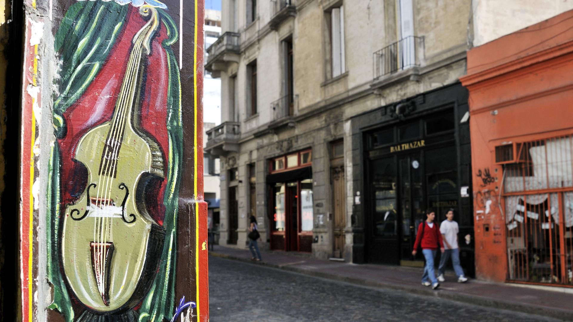 Zwei Menschen gehen im historischer Stadtteil San Telmo in Buenos Aires an einer Hauswand vorbei, die mit einer Geige bemalt ist und dabei von einem grünen Vorhang umgeben ist.