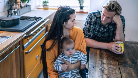 Ein junges Elternpaar sitzt an einem Tisch und schaut sich nachdenklich an. Sie hält einen Säugling in den Armen.