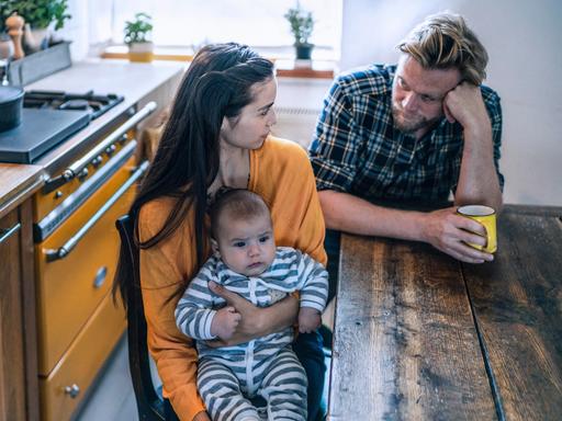 Ein junges Elternpaar sitzt an einem Tisch und schaut sich nachdenklich an. Sie hält einen Säugling in den Armen.