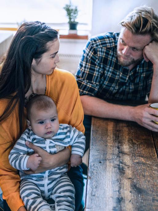Ein junges Elternpaar sitzt an einem Tisch und schaut sich nachdenklich an. Sie hält einen Säugling in den Armen.