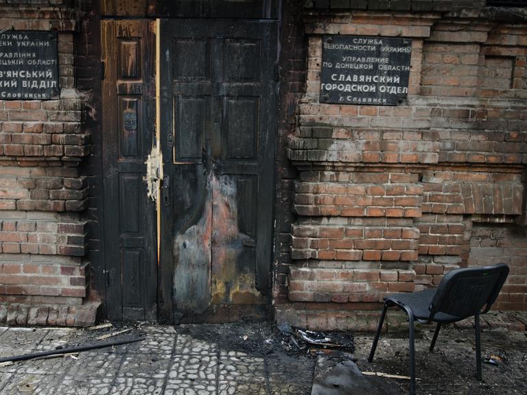 Eine verbrannte Haustür in einem kaputten Haus. Davor steht ein Stuhl.