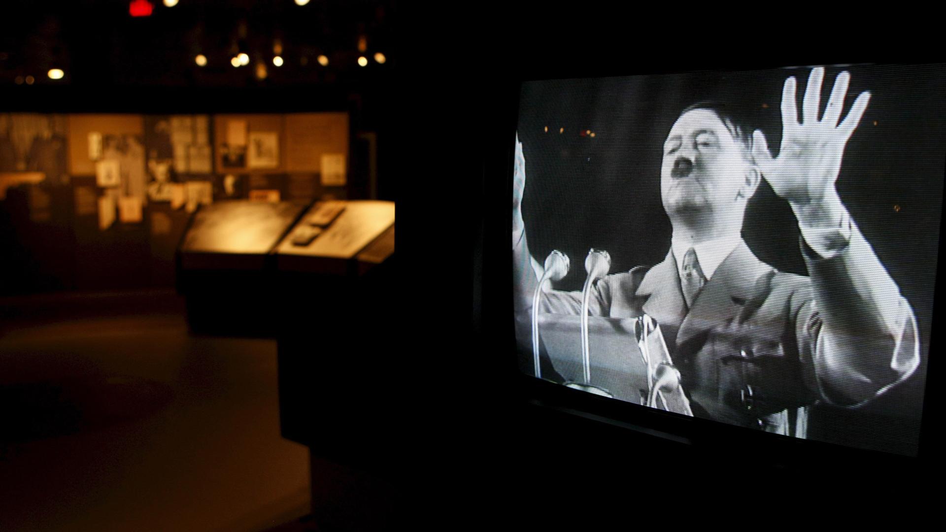 Ein Video zeigt in einem Museum einen Fernseher auf dem Adolf Hitler eine Rede hält. Sein linker Arm ist in die Luft gehoben. Er steht an einem Pult. Vor ihm sind Mikrofone angebracht. 