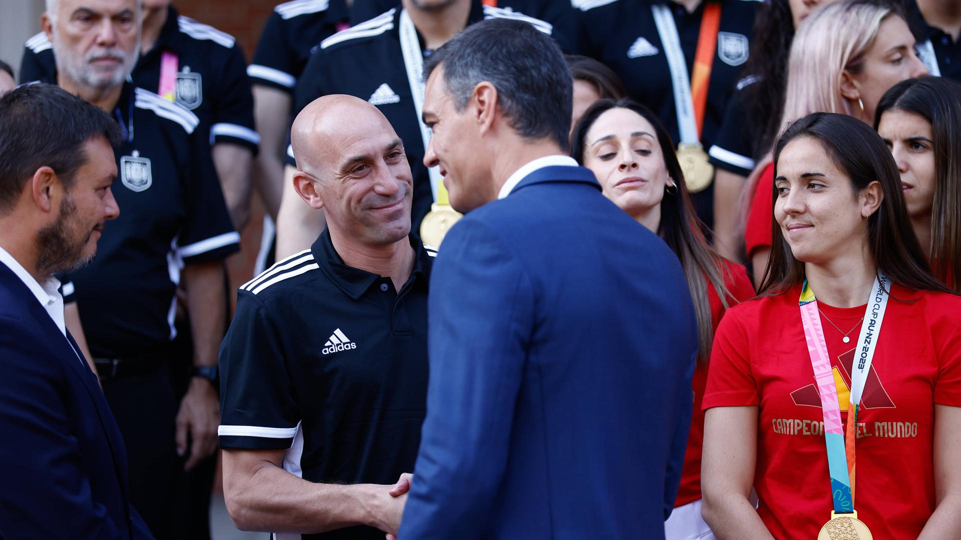 Spaniens Ministerpräsident schüttelt dem Fußball-Verbandspräsidenten Rubiales die Hand, im Hintergrund stehen Spielerinnen.