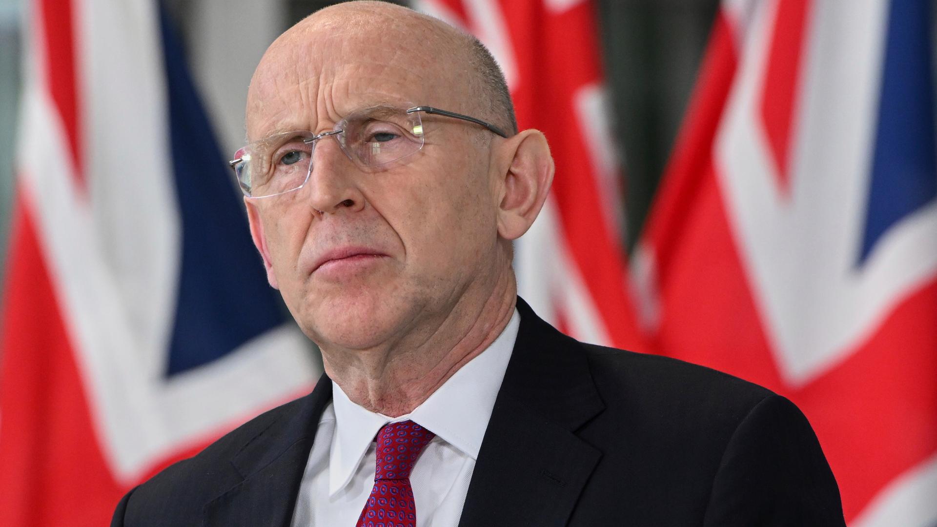 Der britische Verteidigungsminister John Healey in Anzug und Krawatte. Er trägt eine Brille. Im Hintergrund ist die britische Fahne zu sehen.