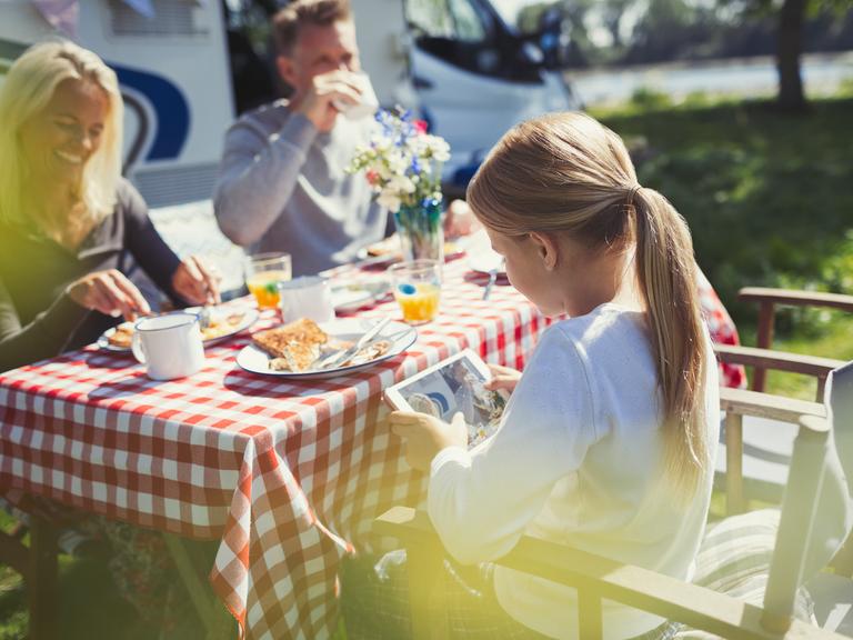 Ein Mädchen schaut auf das Display eines Tablets, während sie draußen, mit Ihrer Familie beim Camping frühstückt.
