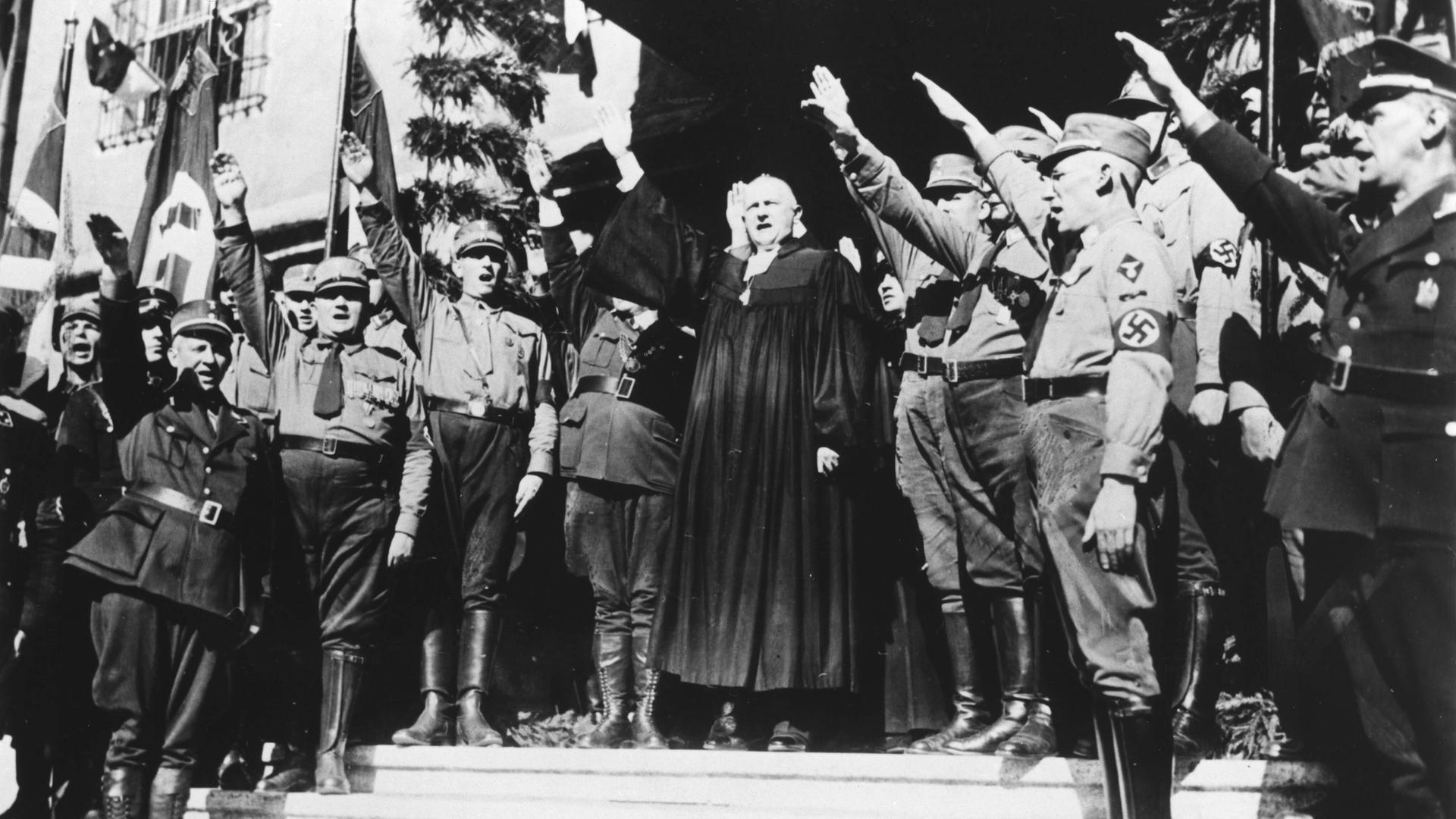 Schwarzweißfoto zeigt einen Mann in Talar, den Hitlergruß zeigend vor einem Spalier von SA-Uniformierten mit erhobenem rechten Armen.