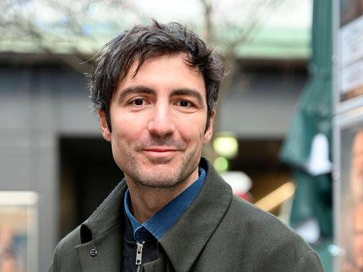 İlker Çatak trägt einen grauen Mantel und posiert im Freien für ein Porträt.