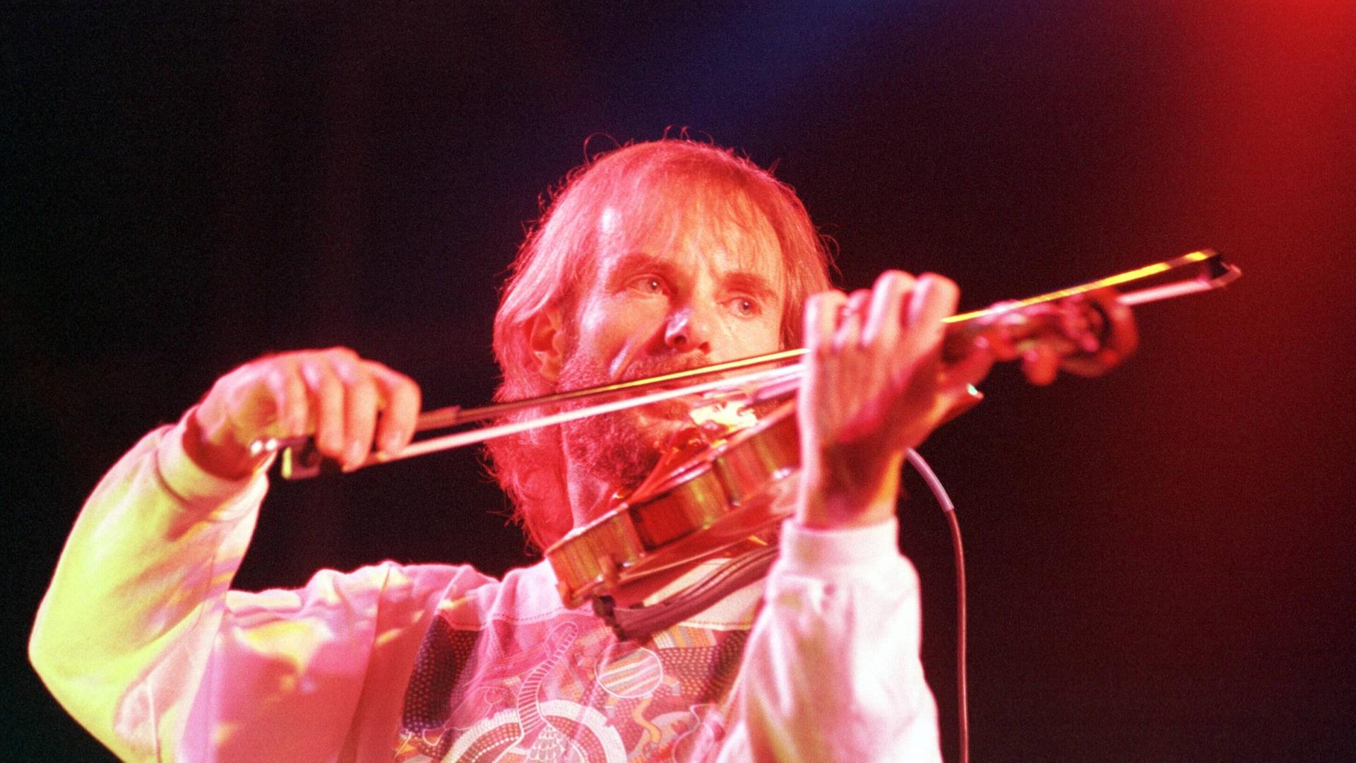 Ein bärtiger Mann spielt, angestrahlt von Bühnenlicht, Geige. 