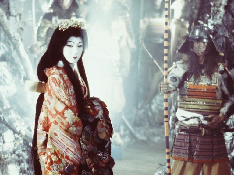 Eine weiß geschminkte japanische Frau in traditionellem Kostüm in einer fantasy-artig anmutenden Umgebung