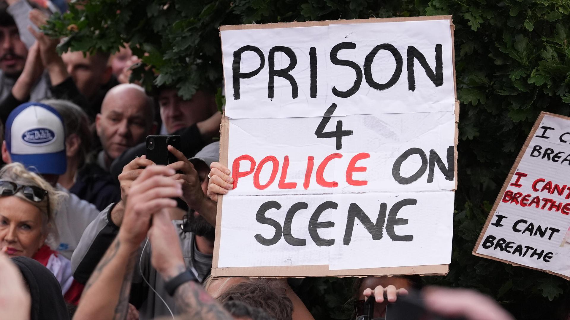 Das Bild zeigt eine Demonstration. Menschen halten ein Schild hoch. Darauf steht: "Prison for police on scene" Das Bild zeigt eine Demonstration. Menschen halten ein Schild hoch. Darauf steht: "Prison for police on scene"