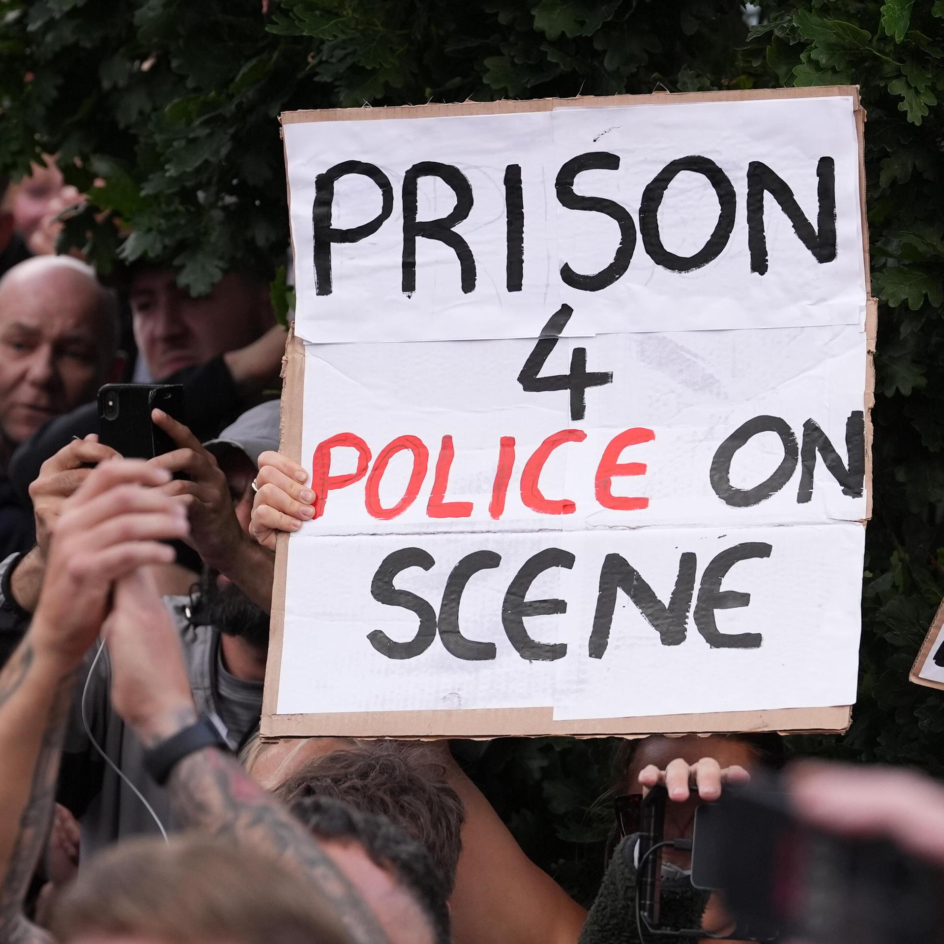 Das Bild zeigt eine Demonstration. Menschen halten ein Schild hoch. Darauf steht: "Prison fpr policen on scene"