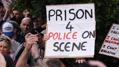 Das Bild zeigt eine Demonstration. Menschen halten ein Schild hoch. Darauf steht: "Prison for police on scene"