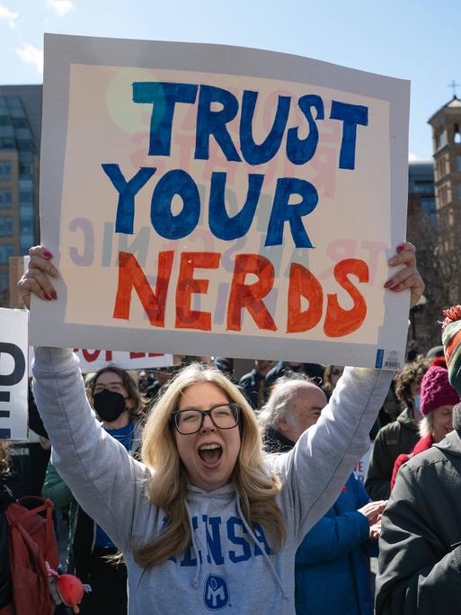Menschen auf einer Demonstration, im Vordergrund steht eine Frau und hält ein Schild mit der Aufschrift "Trust Your Nerds" Menschen auf einer Demonstration, im Vordergrund steht eine Frau und hält ein Schild mit der Aufschrift "Trust Your Nerds"