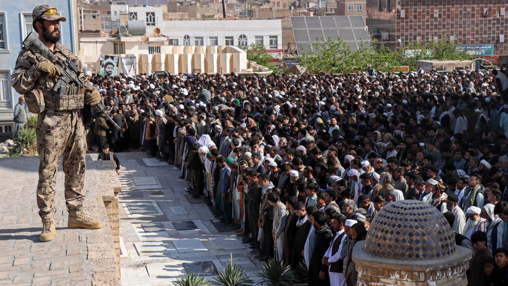 Ein Sicherheitsbeamter der Taliban steht Wache im Jahr 2026, während Männer in Herat/Afganistan ein Gebet sprechen.