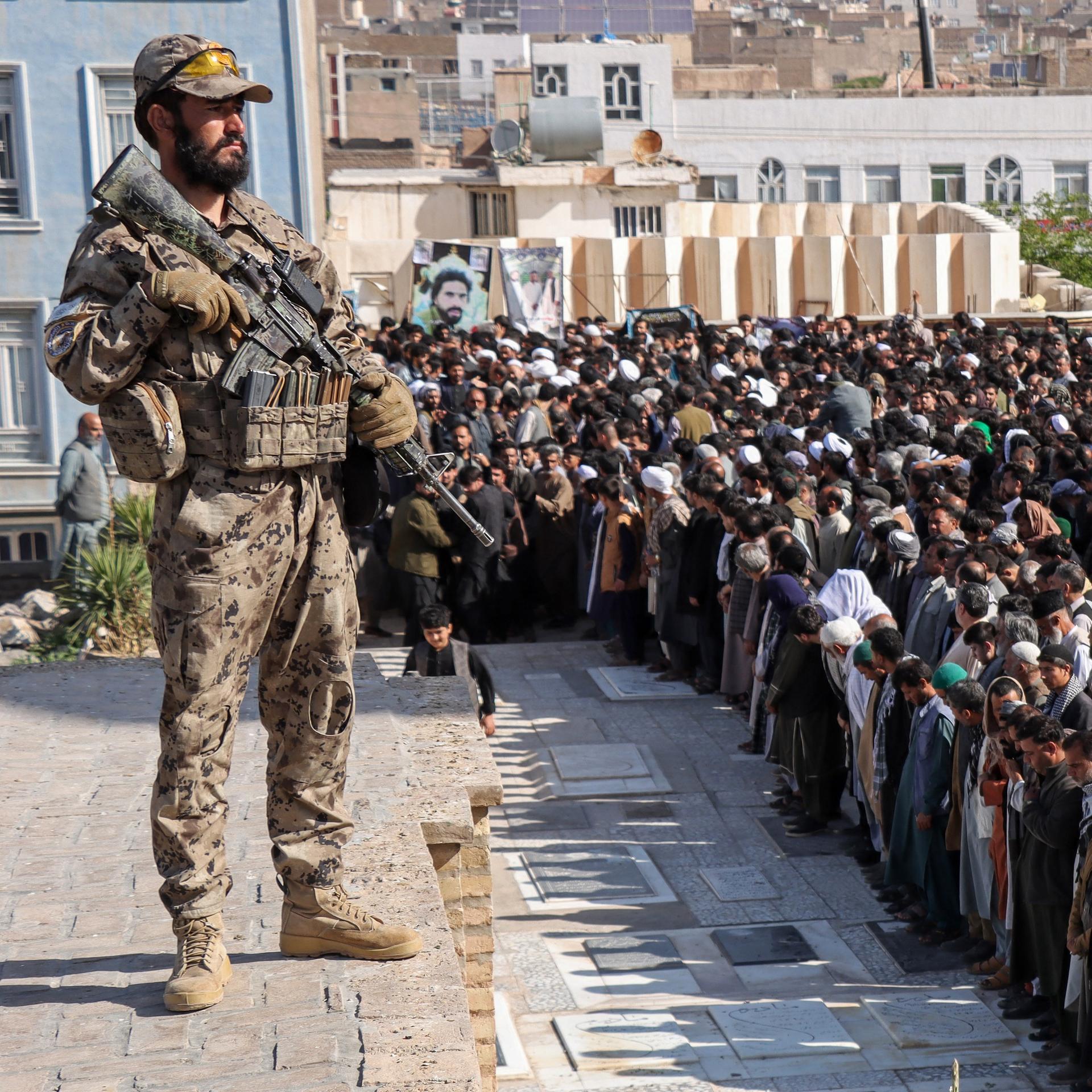 Ein Sicherheitsbeamter der Taliban steht Wache im Jahr 2026, während Männer in Herat/Afganistan ein Gebet sprechen.