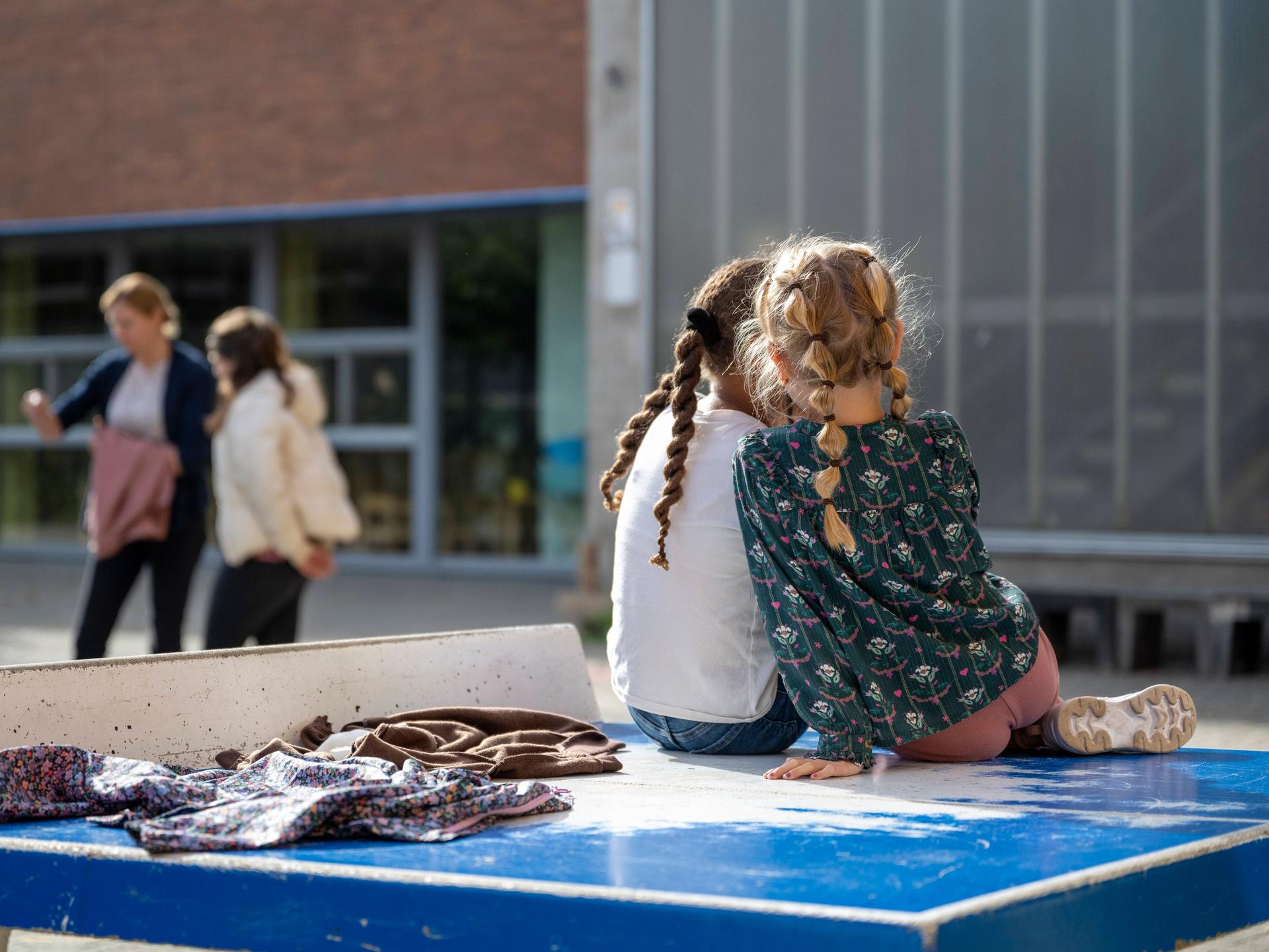 Zwei Schulkinder sitzen mit dem Rücken zum Betrachter auf einer Tischtennisplatte auf einem Schulhof. Im Hintergrund sind in der Unschärfe weitere Personen zu erkennen.