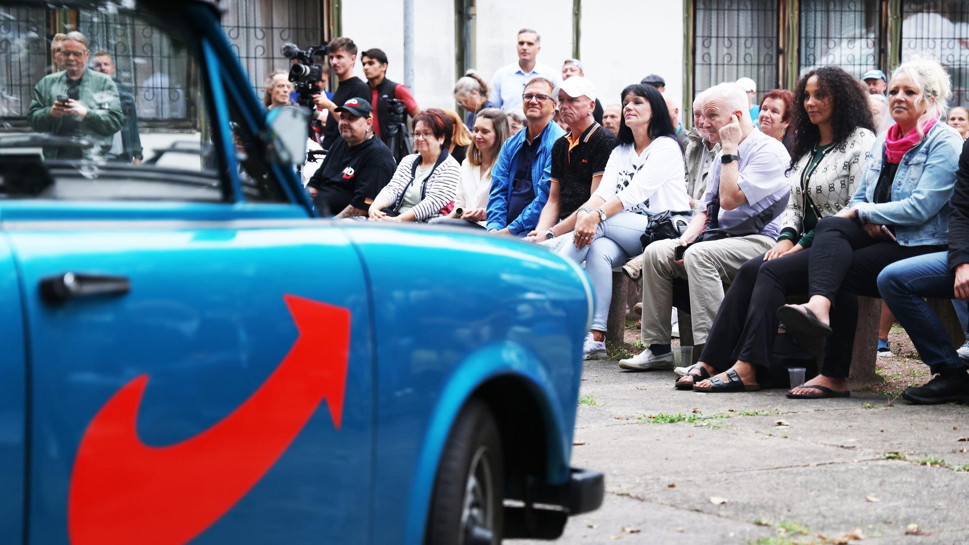 Menschen sitzen in Dessau bei einer Wahlveranstaltung de AfD. Im Vordergrund ist unscharf ein mit AfD Farben und Symbolik lackierter Trabant zu erkennen.