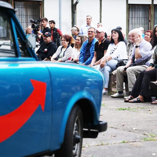 Menschen sitzen in Dessau bei einer Wahlveranstaltung de AfD. Im Vordergrund ist unscharf ein mit AfD Farben und Symbolik lackierter Trabant zu erkennen.