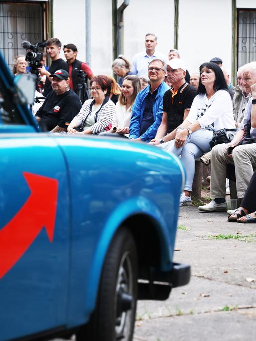 Menschen sitzen in Dessau bei einer Wahlveranstaltung de AfD. Im Vordergrund ist unscharf ein mit AfD Farben und Symbolik lackierter Trabant zu erkennen.