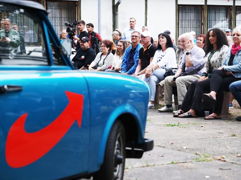 Menschen sitzen in Dessau bei einer Wahlveranstaltung de AfD. Im Vordergrund ist unscharf ein mit AfD Farben und Symbolik lackierter Trabant zu erkennen.