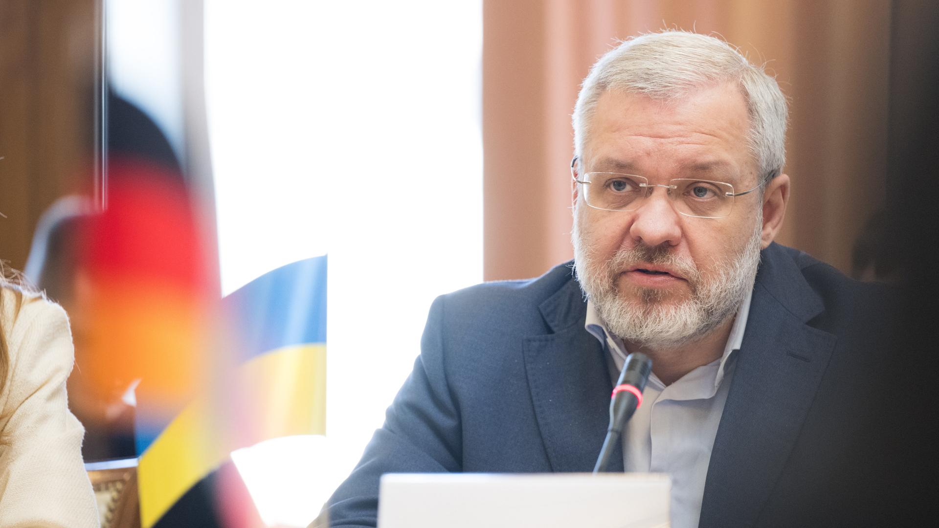 Das Bild zeigt den Justizminister der Ukraine, Herman Haluschtschenko. Er trägt einen dunklen Anzug und sitzt hinter einem Konferenztisch. Dabei blickt er konzentriert nach vorne. Im Hintergrund sind die deutsche und die ukrainische Flagge zu erkennen.