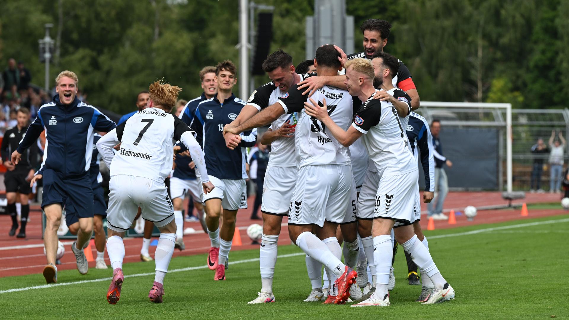 Die Spieler in den weißen Trikots bilden eine jubelnde Spielertraube auf dem Sportplatz. Ersatzspieler in blauen Trainingsjacken kommen dazu. 