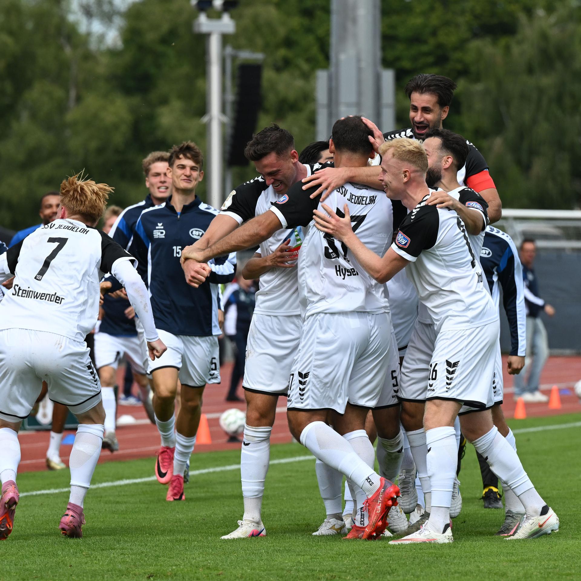 Die Spieler in den weißen Trikots bilden eine jubelnde Spielertraube auf dem Sportplatz. Ersatzspieler in blauen Trainingsjacken kommen dazu. 