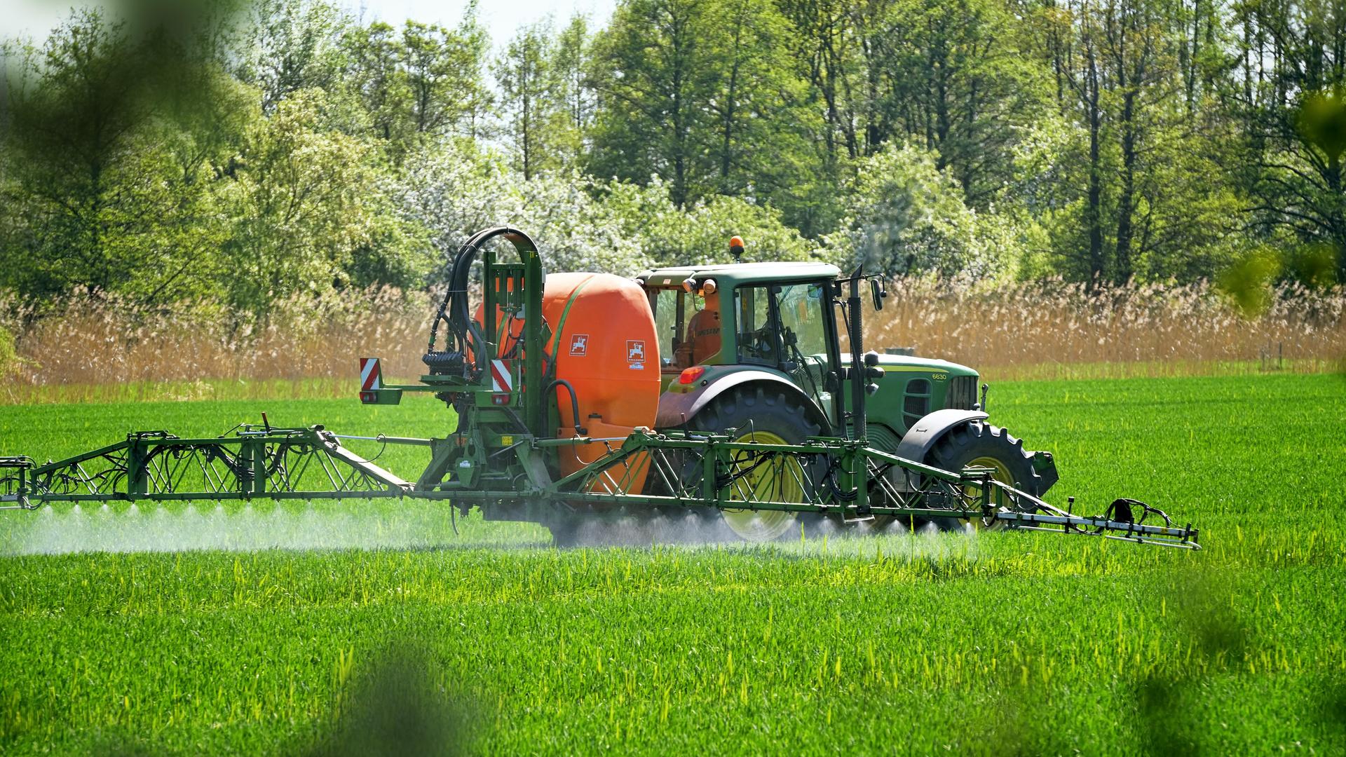 Ein grüner Traktor fährt über einen Acker und versprüht aus einem orangen Tank Pflanzenschutzmittel. Im Hintergrund ist ein Laubwald.