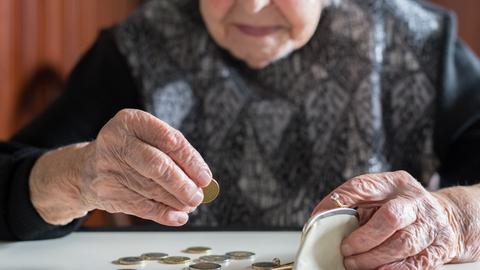 Eine ältere Frau, deren Gesicht durch Anschnitt und Unschärfe nicht erkenntlich ist, sitzt am Tisch und zählt mit faltigen Händen die Münzen aus Ihrer Geldbörse.