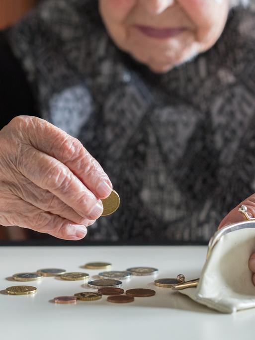 Eine ältere Frau, deren Gesicht durch Anschnitt und Unschärfe nicht erkenntlich ist, sitzt am Tisch und zählt mit faltigen Händen die Münzen aus Ihrer Geldbörse. (Symbolfoto)