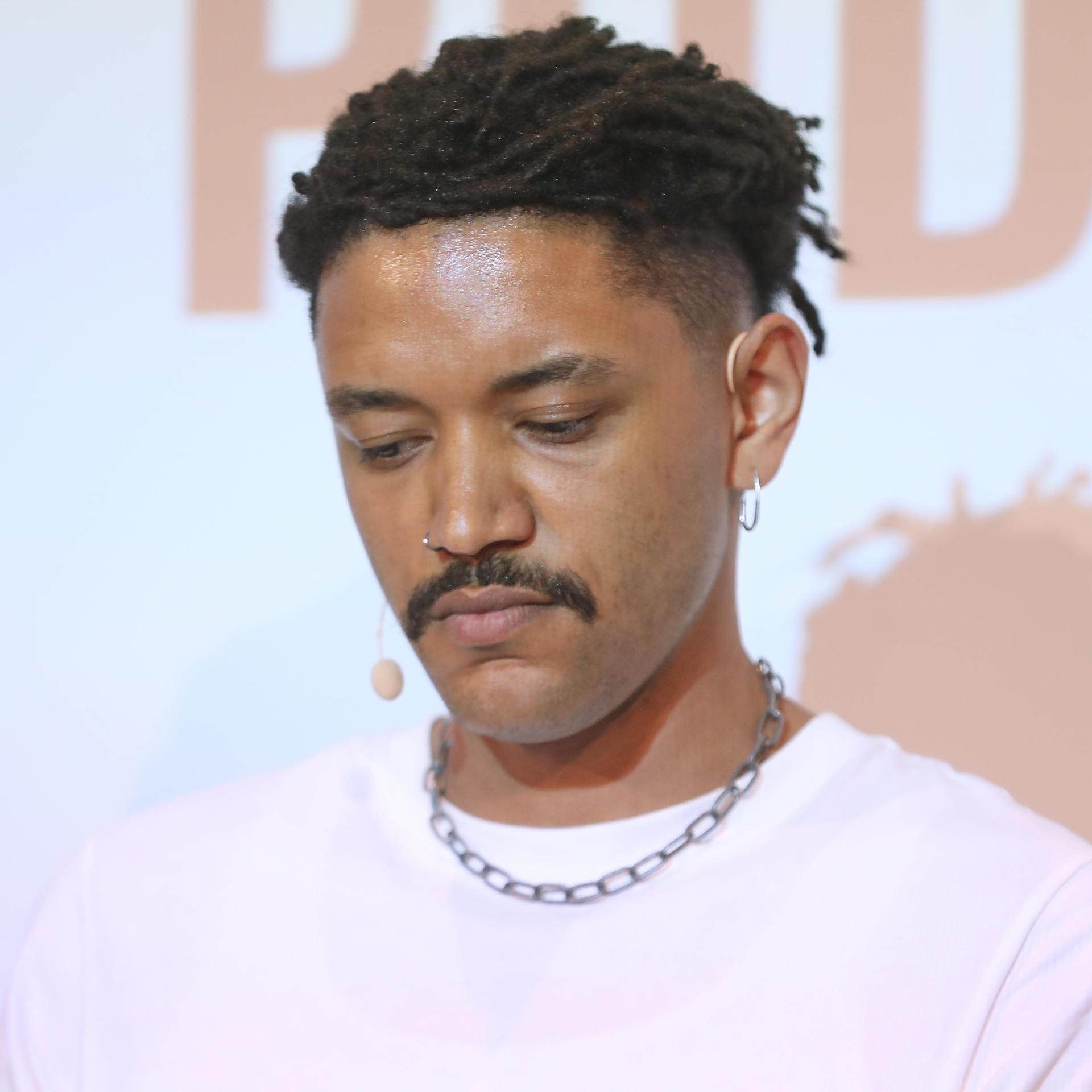 Der Journalist Don Pablo Mulemba blickt nach unten. Er trägt einen Schnurrbart und hat dunkle Haare, ein rosa T-Shirt mit einer Kette.  