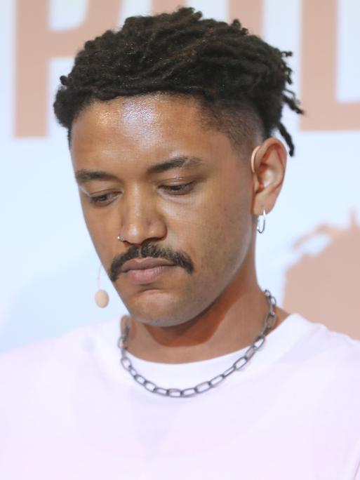 Der Journalist Don Pablo Mulemba blickt nach unten. Er trägt einen Schnurrbart und hat dunkle Haare, ein rosa T-Shirt mit einer Kette. Der Journalist Don Pablo Mulemba blickt nach unten. Er trägt einen Schnurrbart und hat dunkle Haare, ein rosa T-Shirt mit einer Kette.