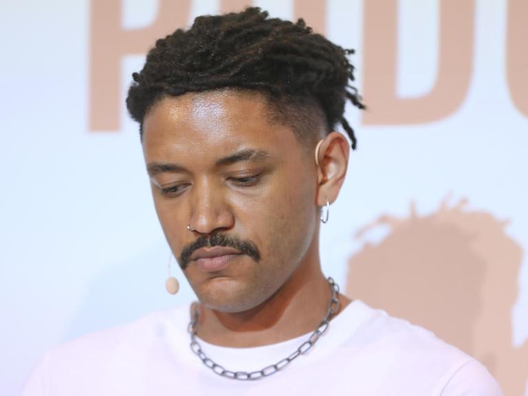 Der Journalist Don Pablo Mulemba blickt nach unten. Er trägt einen Schnurrbart und hat dunkle Haare, ein rosa T-Shirt mit einer Kette. Der Journalist Don Pablo Mulemba blickt nach unten. Er trägt einen Schnurrbart und hat dunkle Haare, ein rosa T-Shirt mit einer Kette.