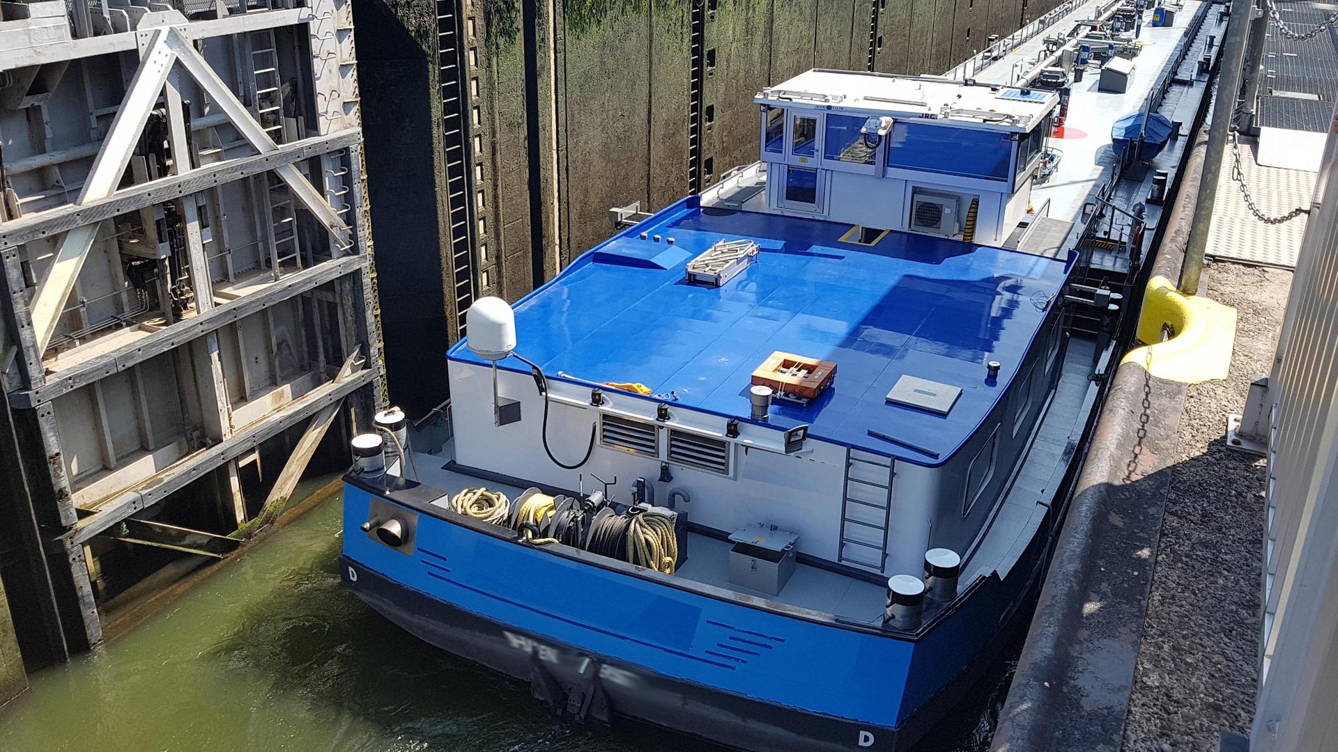 Ein Schifffährt in eine Schleusenkammer ein. Das Schiff ist teilweise blau, die Schleuse ist teilweise grau. Die Hänge des Ufers sind steil.