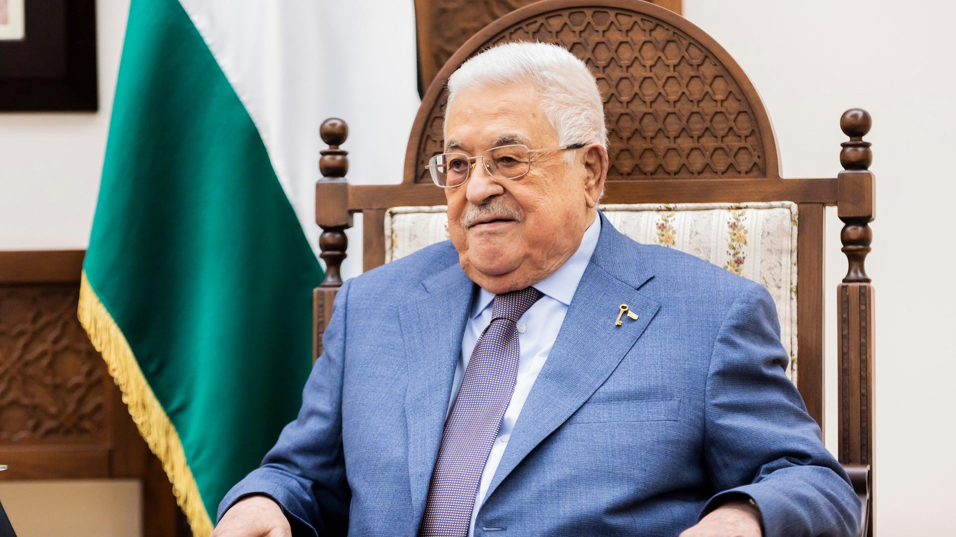 Abbas im blauen Jackett sitzt in einem Stuhl und blickt ernst. naben ihm eine palästineneische Fahne.