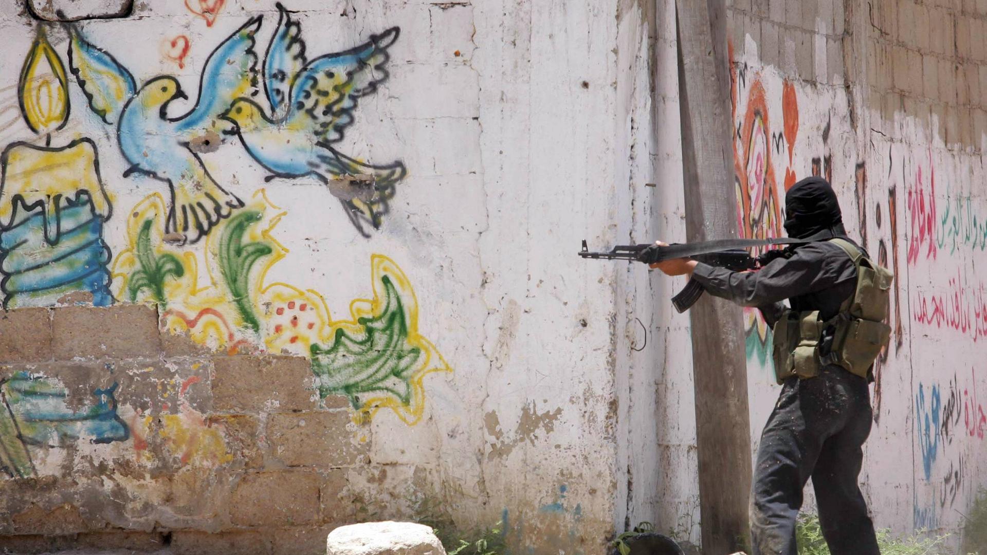 Ein Mitglied der Al-Qassam Brigade zielt mit einem Gewehr an einer Mauer in Gaza Stadt. Auf der Mauer ist ein Graffiti mit bunten Tauben als Symbol für Frieden zu sehen.